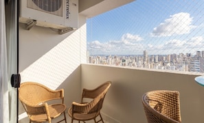 A balcony with two wicker chairs, a small table, and a city view through a chain-link fence.