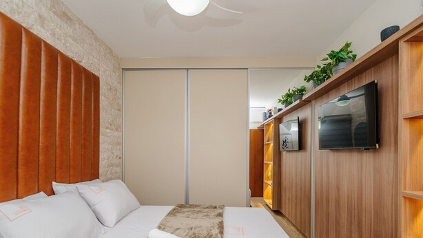 A modern bedroom with a bed, wooden headboard, a TV, and a shelf with plants. - Apt in Cond. w/ Pool in Goiania Bli1801 (Goiânia)