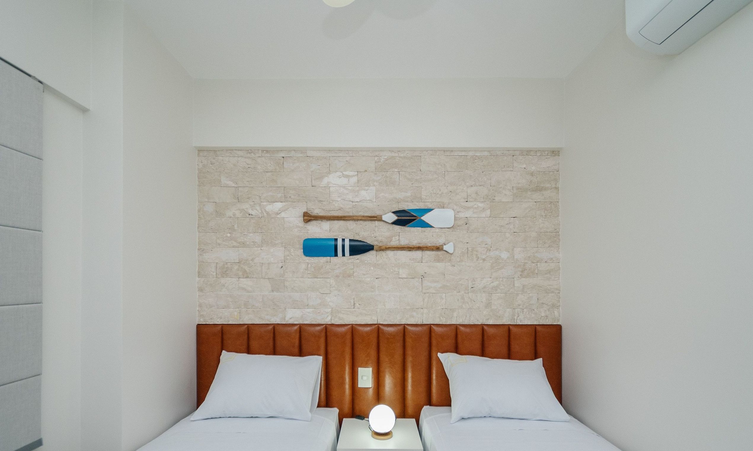 Un dormitorio con pared de ladrillo, dos camas con ropa de cama blanca y dos palas colgadas en la pared.