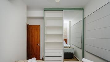 A room with a wooden door, a white wardrobe with horizontal handles, a bed with a white mattress and brown headboard, and a grey padded wall.