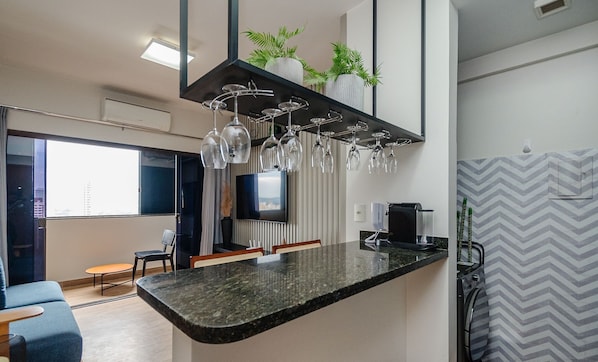 A modern kitchen with a granite countertop, hanging wine glasses, a built-in TV, and a coffee machine.