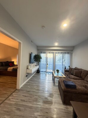 A modern living room with a sofa, a coffee table, and a TV. There is a sliding glass door leading to a balcony.