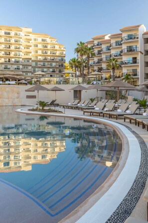 Piscina con sillas de descanso y sombrillas, rodeada de edificios de varios pisos con balcones.