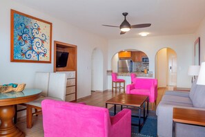 A modern living room with a round coffee table, two pink armchairs, a grey sofa, and a wooden dining table. A ceiling fan and a TV are mounted on the wall. - Luxury Resort with 1 bedroom and 1.5 bathrooms in beautiful Cabo San Lucas (Cabo San Lucas)
