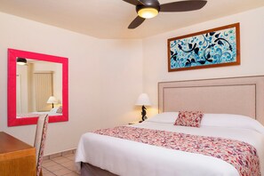 Habitación de hotel con cama, escritorio, silla, ventilador de techo, lámpara de pared y una obra de arte enmarcada.