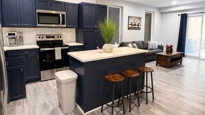 A modern kitchen with dark blue cabinets, stainless steel appliances, a central island with stools, and a large window providing natural light. - Old Town Manassas Sleeps 4 next to bars and restaurants ,  WALK TO VRE (Manassas)