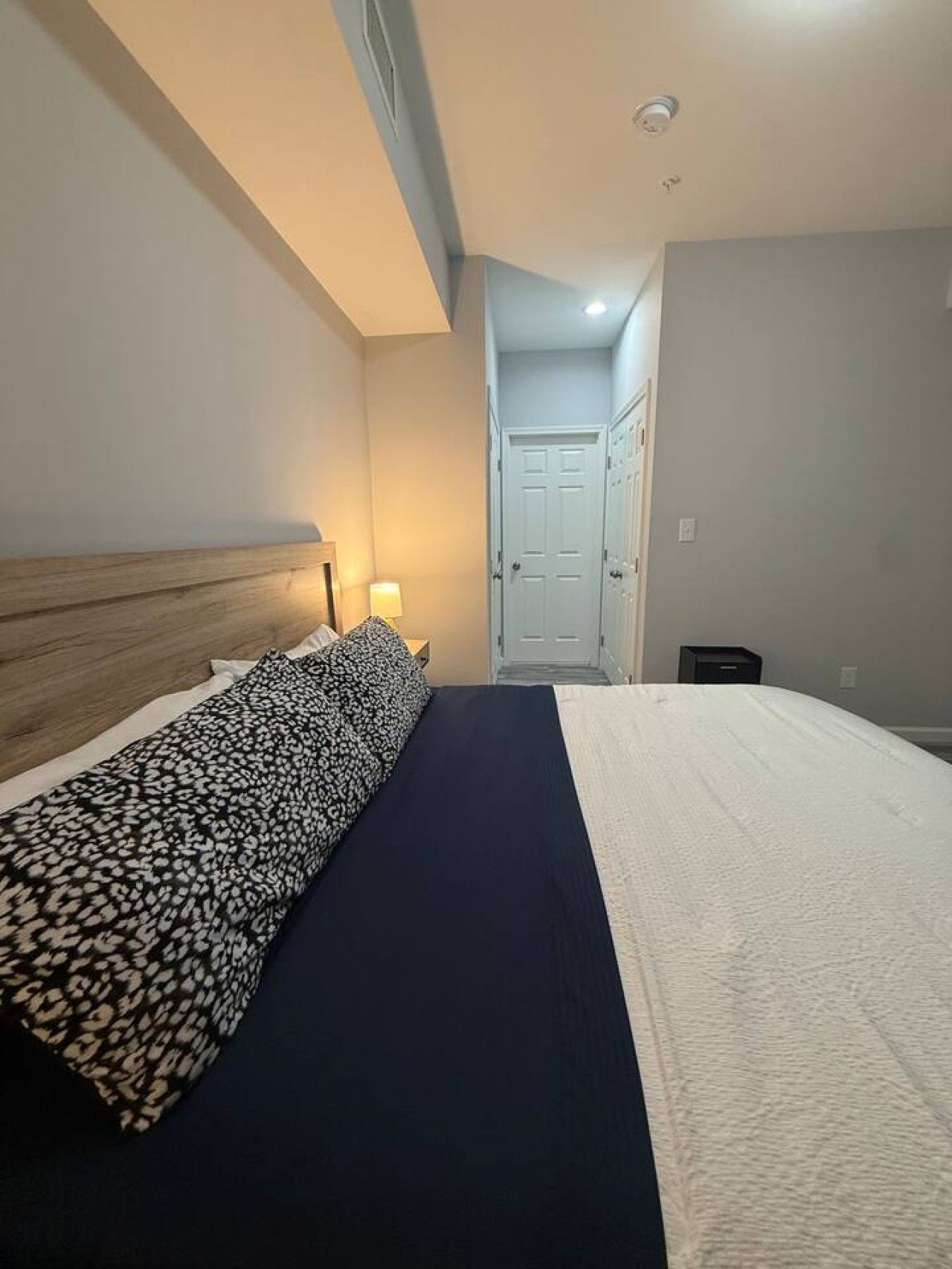 A hotel room with a bed, a nightstand, a lamp, and a door leading to another room.