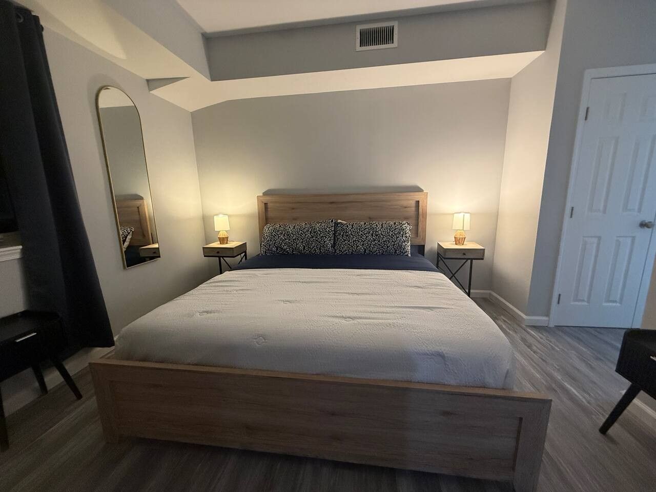 A bedroom with a large bed, two bedside tables with lamps, a mirror, and a door.
