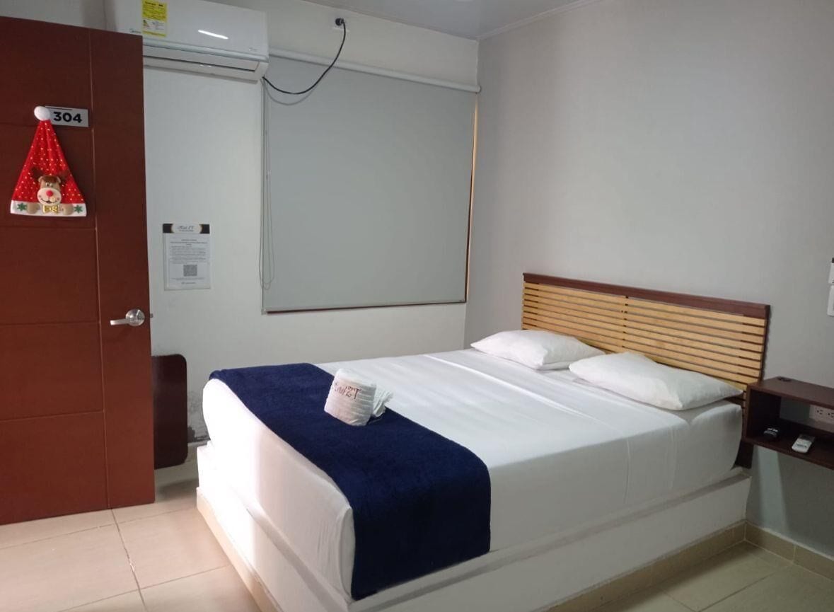 Habitación de hotel con cama, cabecera de madera, mesita de noche, aire acondicionado instalado en la pared y puerta con un cartel de número.
