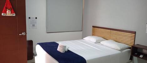 Habitación de hotel con cama, cabecera de madera, mesita de noche, aire acondicionado instalado en la pared y puerta con un cartel de número.