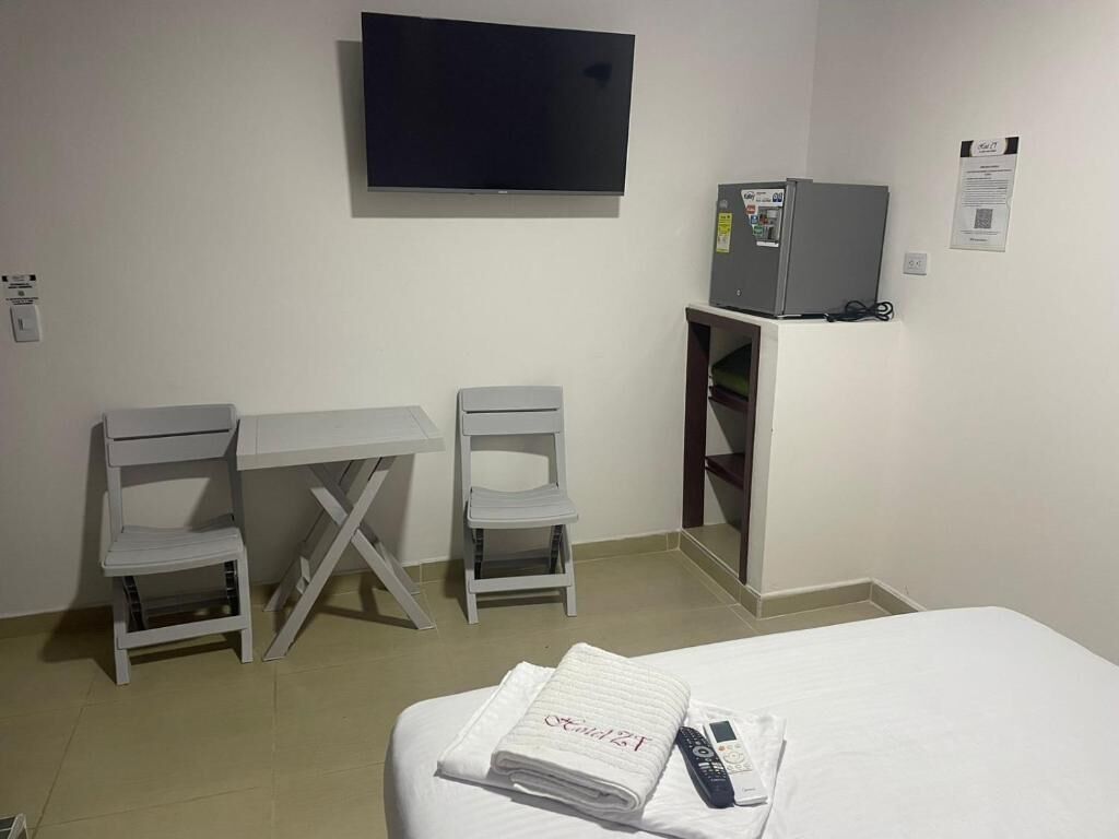 A hotel room with a bed, a desk with two chairs, a television, a refrigerator, and a remote control on the bed.