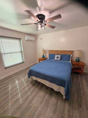 Quarto com cama, duas mesas de cabeceira, ventoinha no teto, janela com persianas e aparelho de ar condicionado.