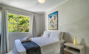 Uma cama bem arrumada com roupas de cama brancas e cinzas, um quadro com balões de ar quente na parede e uma janela com cortinas.
