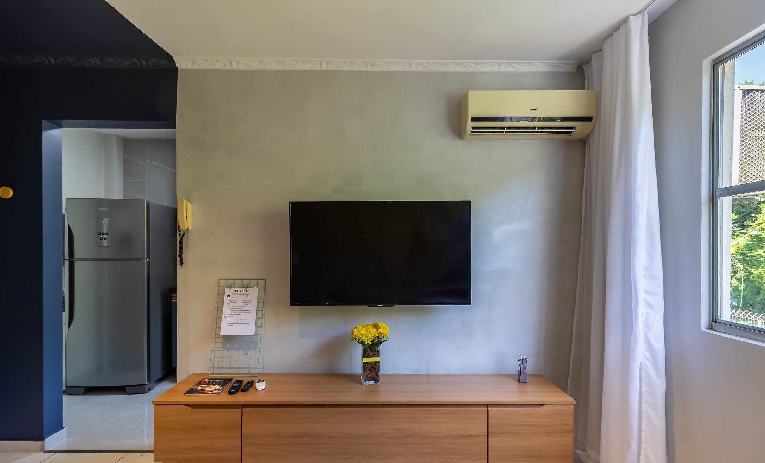 A modern room with a flat-screen TV mounted on the wall, a wooden cabinet with a vase of flowers, a refrigerator, and an air conditioning unit.