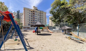 Un parc infantil amb cadenes, tobogan i una caseta de joc amb teulada de palla, envoltat per una tanca i edificis al fons.