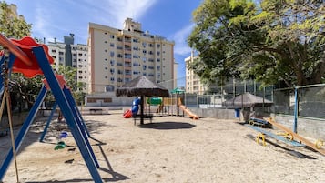 A playground with swings, a slide, and a thatched-roof playhouse, surrounded by a fence and buildings in the background.