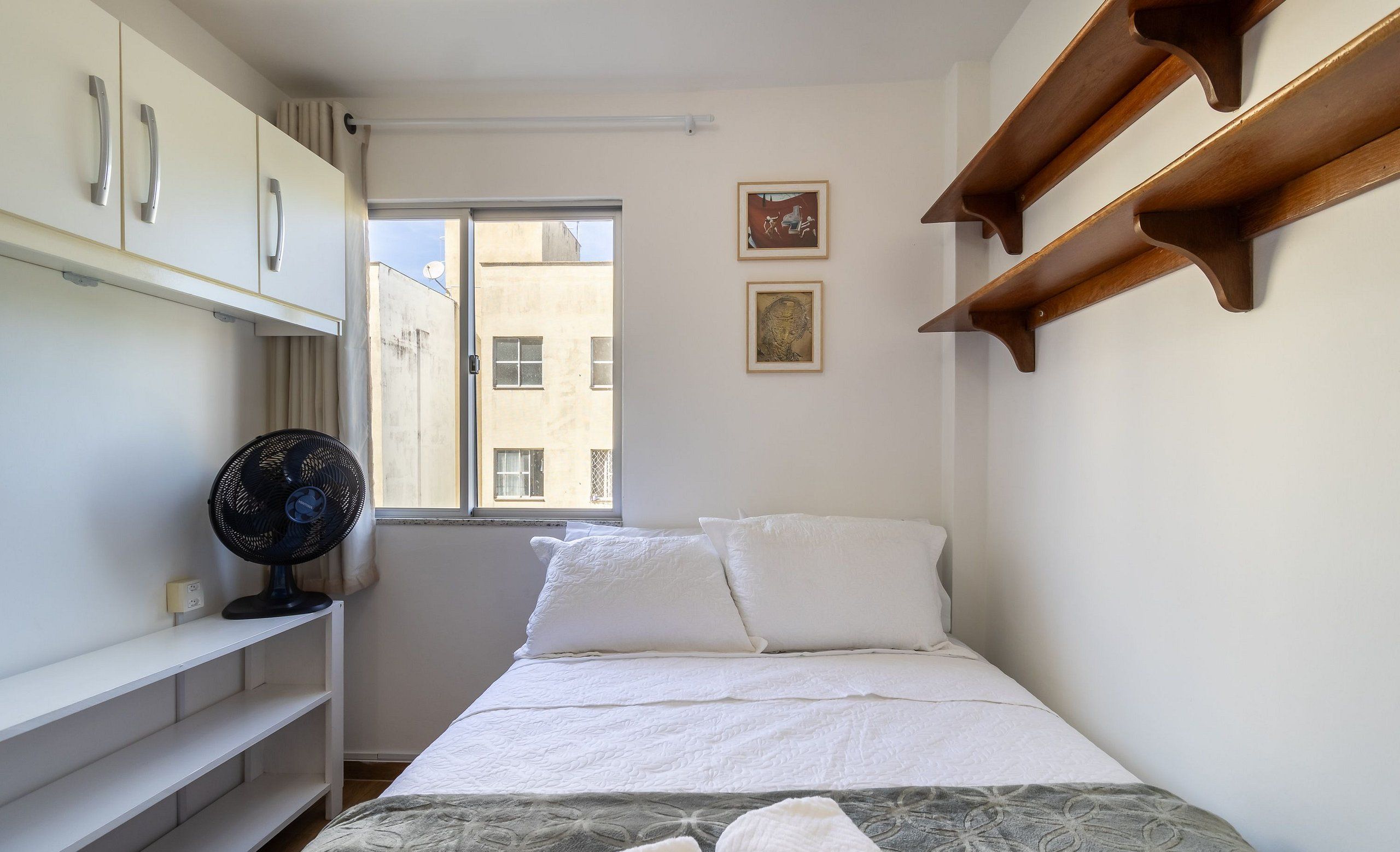 A small, neatly arranged bedroom with a bed, a fan, a window, and a wall-mounted shelf.