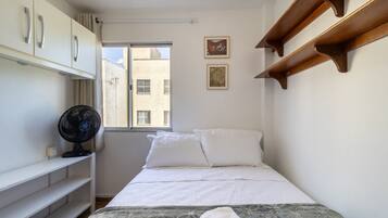 A small, neatly arranged bedroom with a bed, a fan, a window, and a wall-mounted shelf.