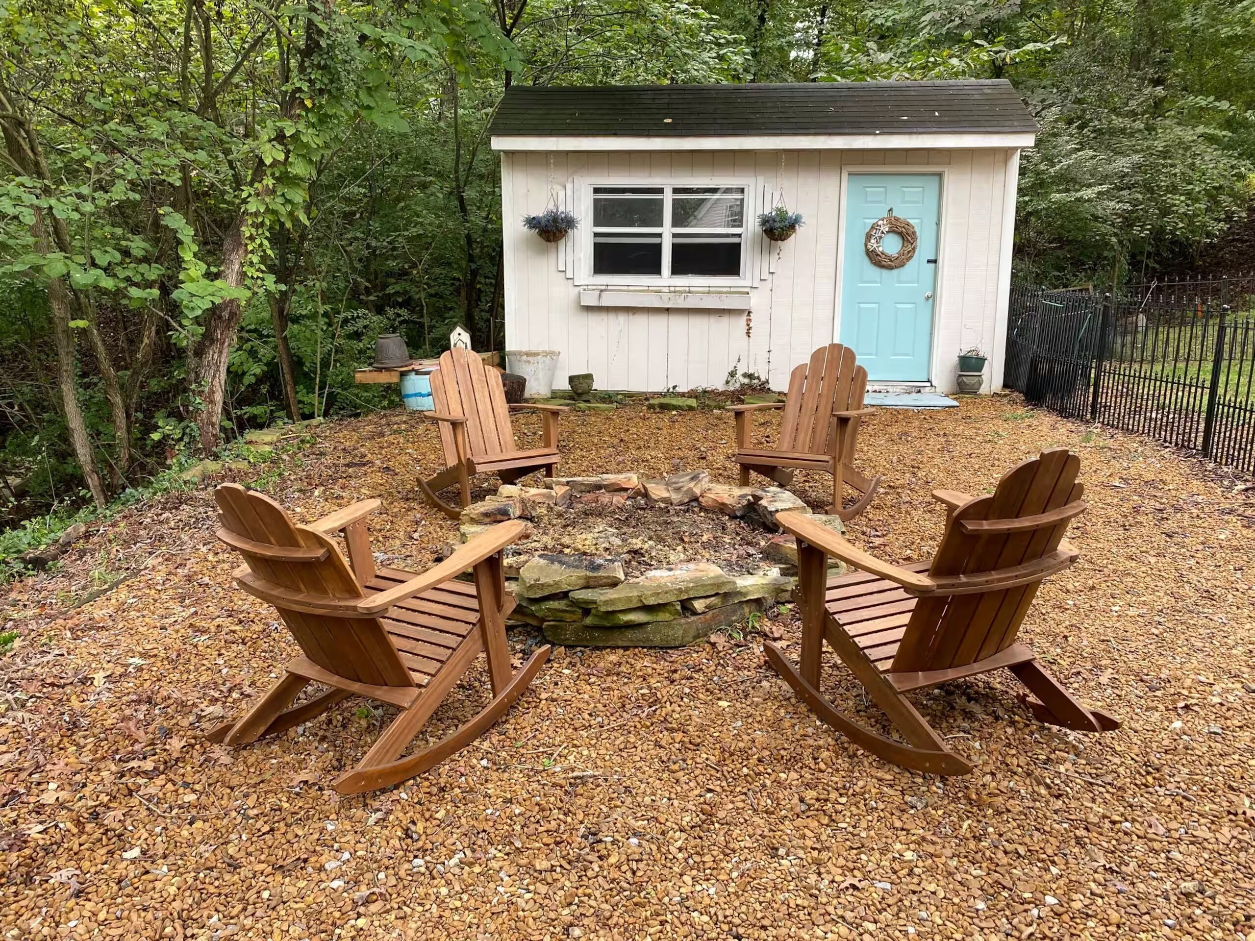 A wooden deck with Adirondack chairs and a fire pit, surrounded by a white building with a blue door, set in a wooded area.