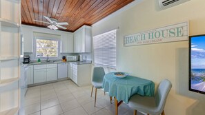 A kitchen with white cabinets, a microwave, and a coffee maker. There's a dining area with a table and chairs, and a sign that reads 'Welcome to our Beach House Anna Maria Island.'