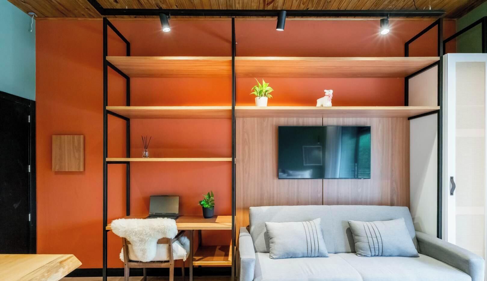 Ein modernes Zimmer mit einem Bett, einem Schreibtisch mit einem Laptop, einem Sessel, einem an der Wand montierten Fernseher und Regalen mit Pflanzen.