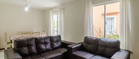 A living room with a dark brown leather sofa, a dining table with chairs, and a window with curtains.