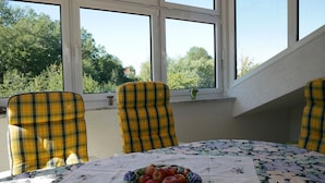 Ein Essbereich mit Tisch, gedeckt für vier Personen, mit einer blumigen Tischdecke und einer Obstschüssel. Die Stühle haben ein Karomuster.