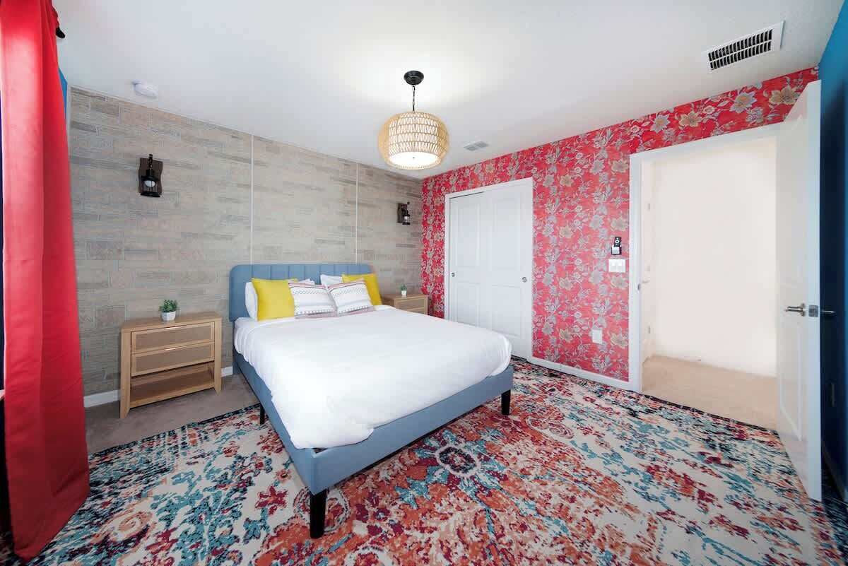 A bedroom with a bed, a nightstand, a patterned rug, a door, and a wall with a floral design.
