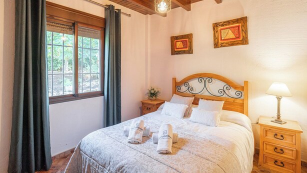 A bedroom with a bed, a nightstand, a lamp, a window with curtains, and two framed pictures on the wall. - Villa Ana Manuela El Gastor by Ruralidays (Cádiz)
