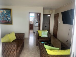 A living room with wicker furniture, a television, and a kitchen in the background. - Adorable 3-bedroom, 2.5 bath apartment with a balcony overlooking the ocean. (Salinas)