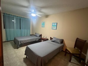 A hotel room with two single beds, a ceiling fan, a small table with a chair, and a window with curtains. - Adorable 3-bedroom, 2.5 bath apartment with a balcony overlooking the ocean. (Salinas)