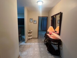 A hotel hallway with a carpeted floor, a wooden console table with a lamp, a mirror, and a small shelf with a decorative item. - Adorable 3-bedroom, 2.5 bath apartment with a balcony overlooking the ocean. (Salinas)