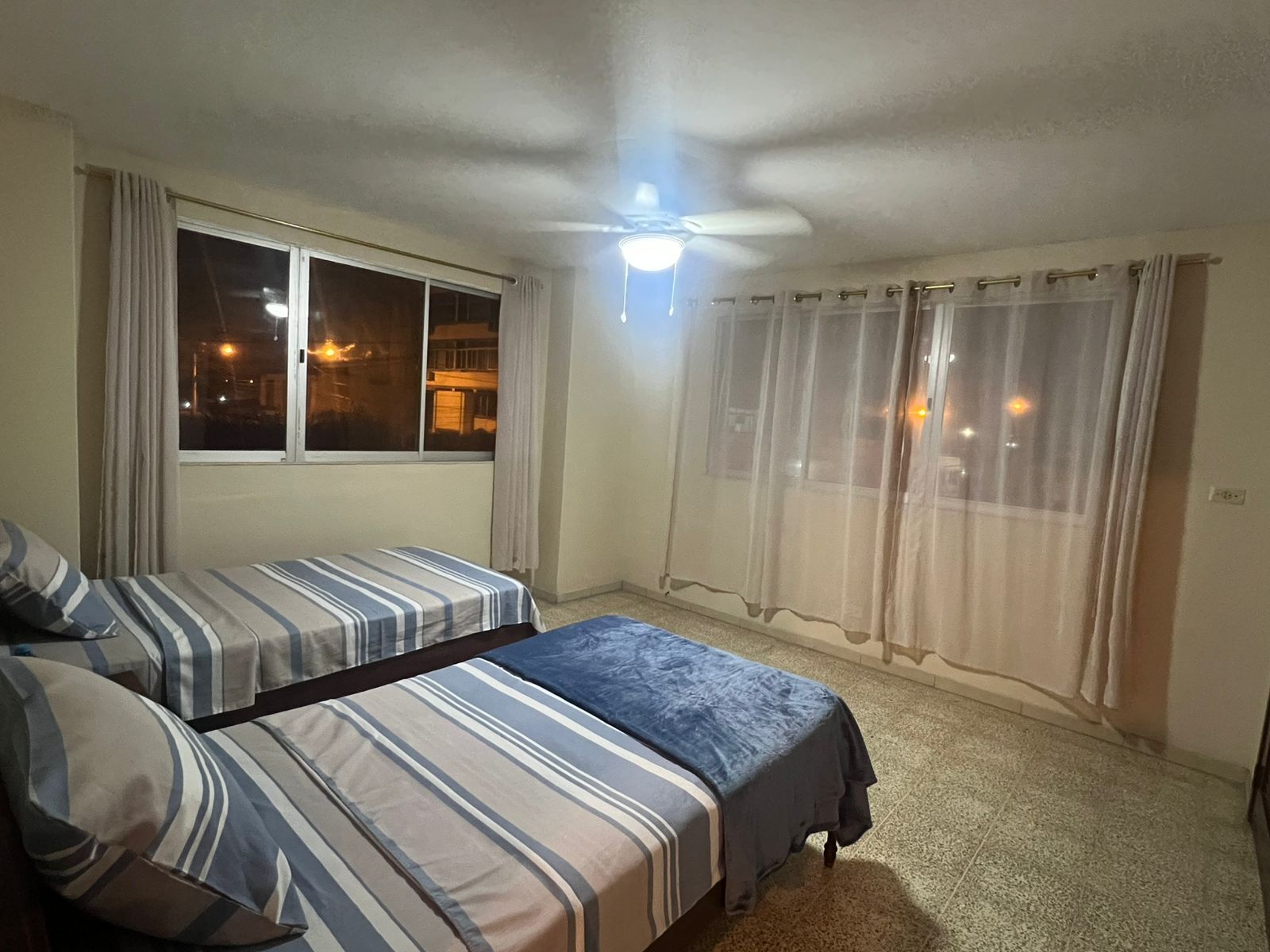 Ein Zimmer mit zwei Betten, einem Deckenventilator, einem Fenster mit Vorhängen und Blick ins Freie bei Nacht.