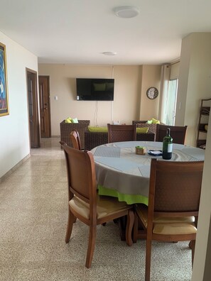 A dining area with a round table, chairs, a television mounted on the wall, and a clock. - Adorable 3-bedroom, 2.5 bath apartment with a balcony overlooking the ocean. (Salinas)