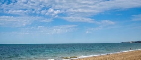 A beach with pebbles, a person lying on a mat, and the sea under a partly cloudy sky.