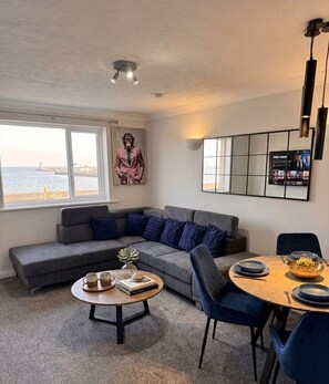 A living room with a grey sofa, a round coffee table, and a dining area with a wooden table and chairs. - Stunning Sea View Flat on The Leas Panoramic Estuary Sunsets, Steps to Beach (Sheerness)