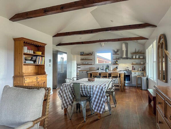 A spacious kitchen with wooden beams, a dining area with a checkered tablecloth, and a cozy seating area with a cushioned chair.