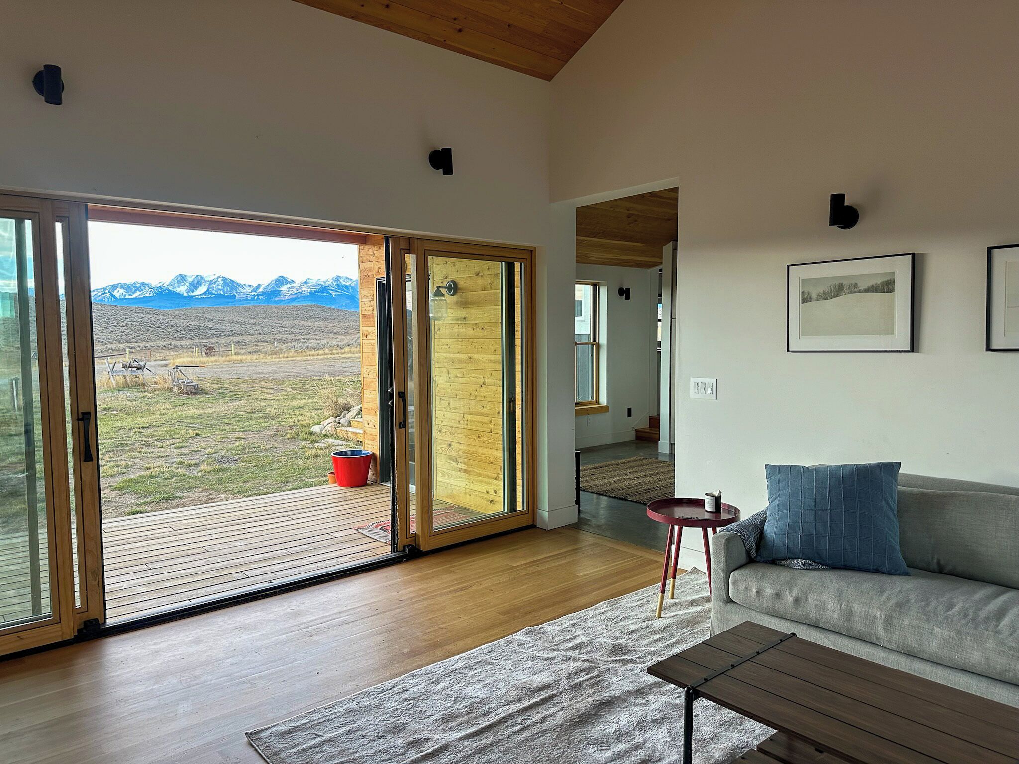 A modern living room with a large sliding glass door leading to a deck, a grey sofa, a small round table, and a wooden coffee table.