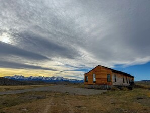 A wooden cabin with a single-story structure, a sloped roof, and a chimney, situated in a rural landscape with mountains in the background. - Private Bison Ranch With Stunning Mountain Views (Wilsall)