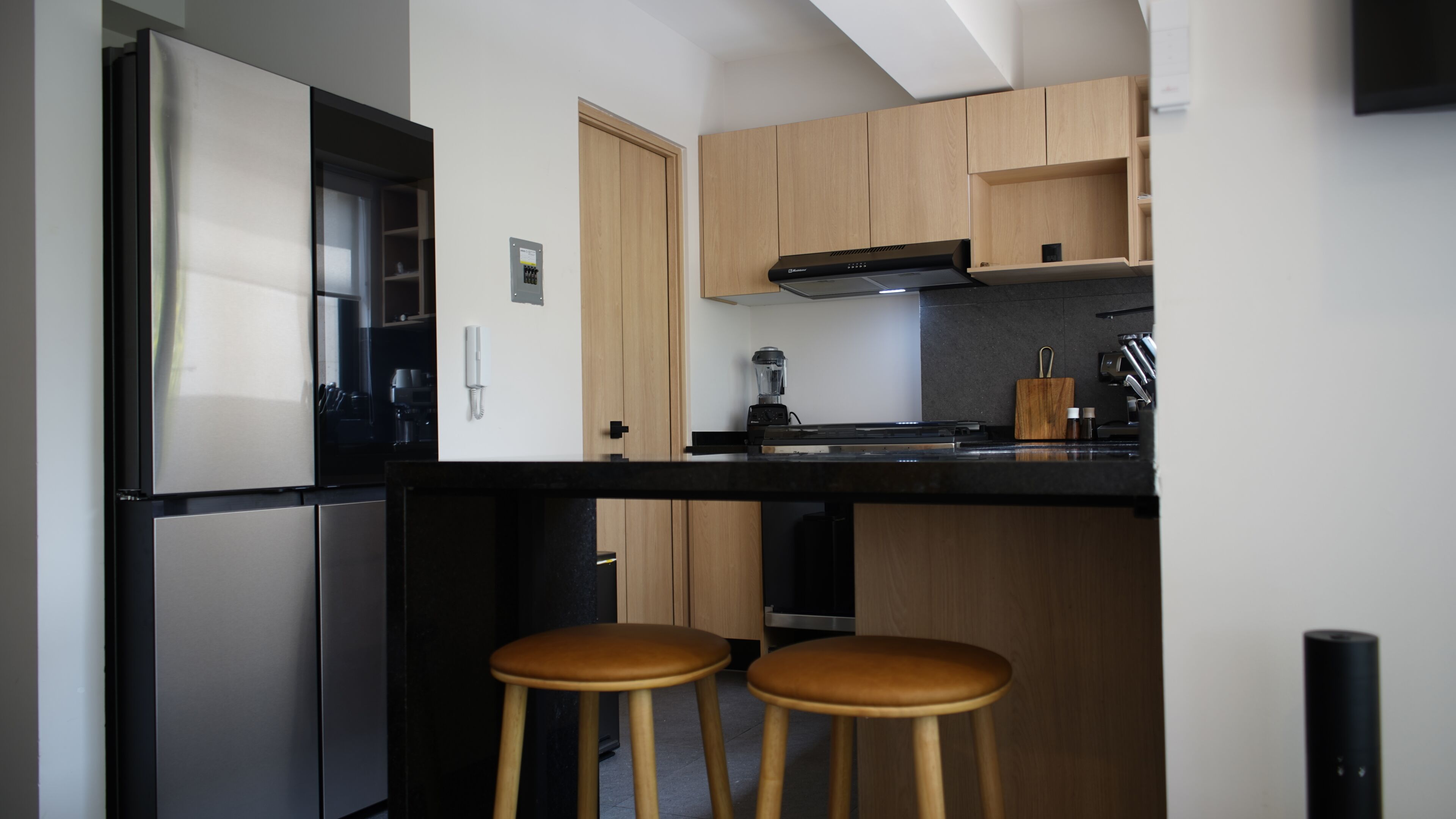 Una cucina moderna con frigorifero integrato, piano cottura e frullatore sul ripiano. Sono presenti due sgabelli di legno e una piccola mensola in legno sopra il bancone.