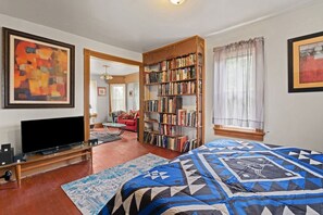 Ein Zimmer mit Fernseher, Bücherregal, rotem Sofa und einem gemusterten Bettüberwurf.