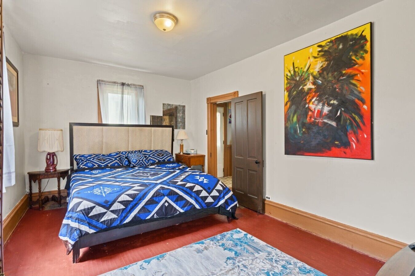Una camera da letto con un letto, un comodino, una lampada, una porta e un quadro appeso al muro.