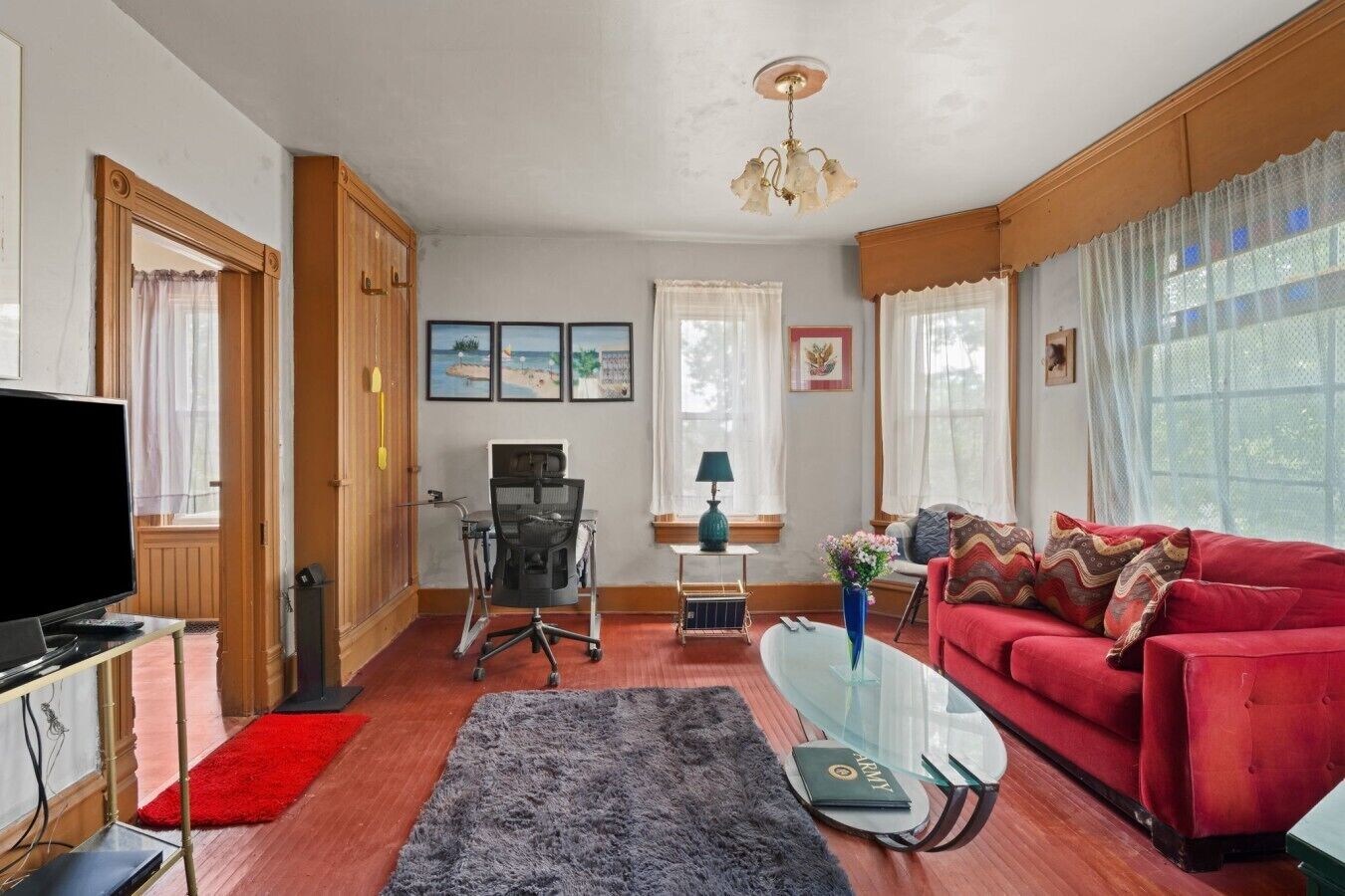 Ett vardagsrum med en röd soffa, ett soffbord i glas, en tv och ett skrivbord med en dator.