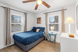 Ein Schlafzimmer mit einem Bett, einem Deckenventilator, einem Nachttisch mit Lampe und einem Fenster mit Vorhängen.