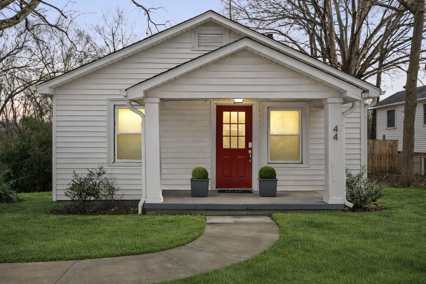 A single-story house with a red front door, two windows on either side, and a small porch with two potted plants.