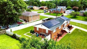Een woonwijk met huizen, waarvan er één een dak met zonnepanelen heeft, omgeven door groen en een heldere lucht.
