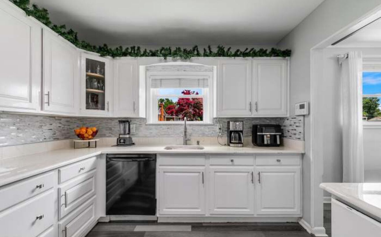 A kitchen with white cabinets, a dishwasher, a coffee maker, and a window with a view of a garden.