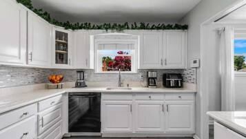 A kitchen with white cabinets, a dishwasher, a coffee maker, and a window with a view of a garden.