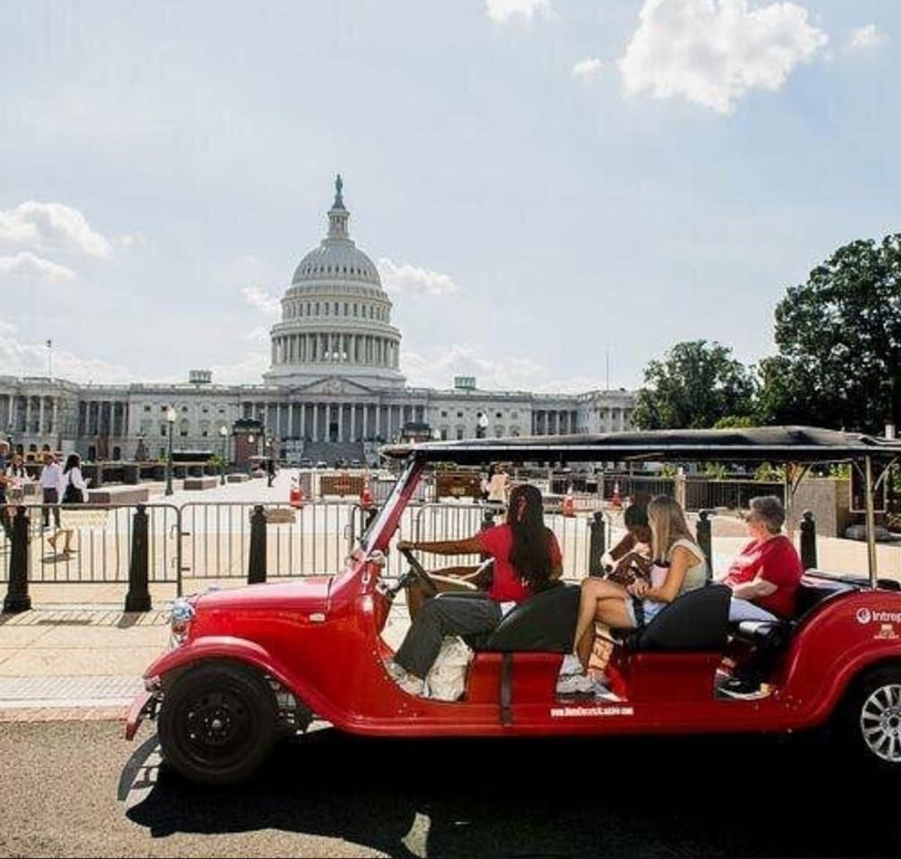 A red vintage car with passengers, parked in front of a large white building with columns.