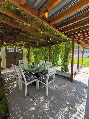 Eine Terrasse im Freien mit einem Glastisch und weißen Stühlen, umgeben von Grünpflanzen und Holzbalken.